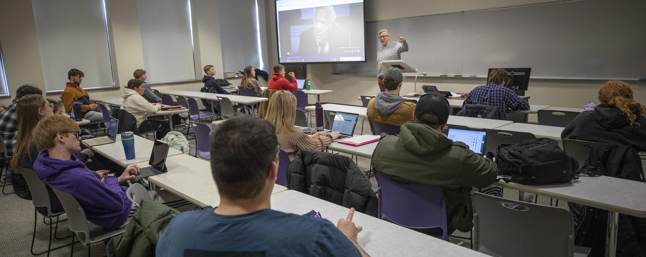 A faculty member points to a whiteboard at the front of class.