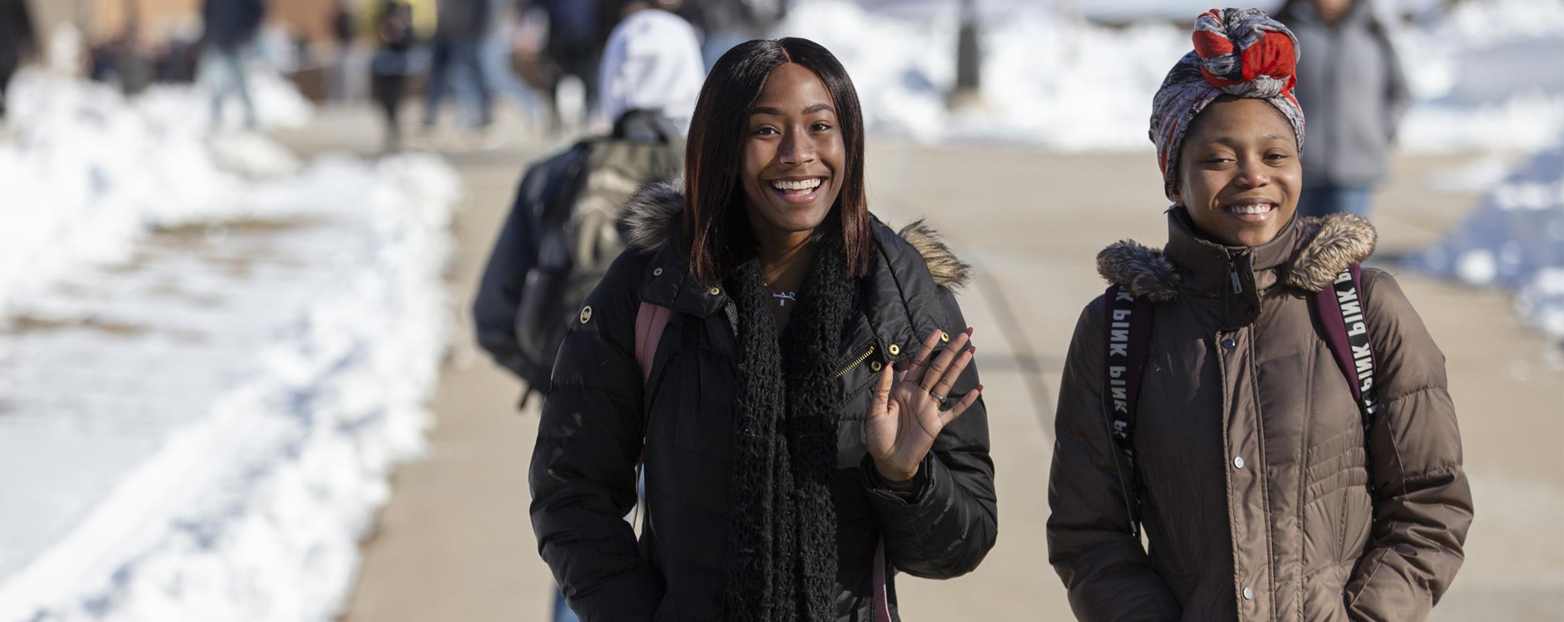 Two students walk outside wearing winter jackets and smile at the camera.